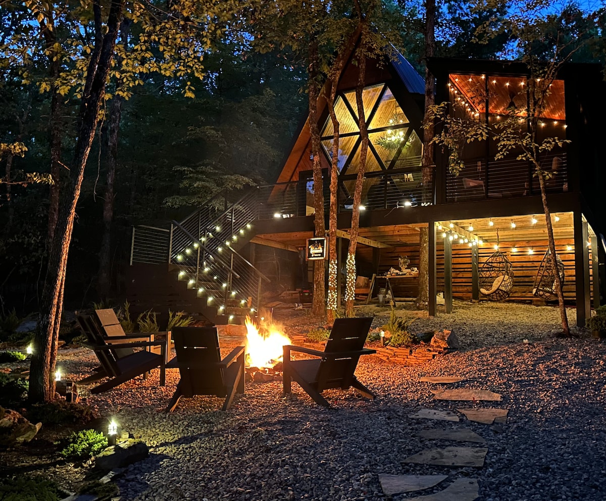 The cabin's exterior is illuminated at night, featuring an inviting fire pit surrounded by comfortable seating. Charming string lights accentuate the architectural design, while stairs lead to a spacious deck with additional seating, nestled within a tranquil forested area.
