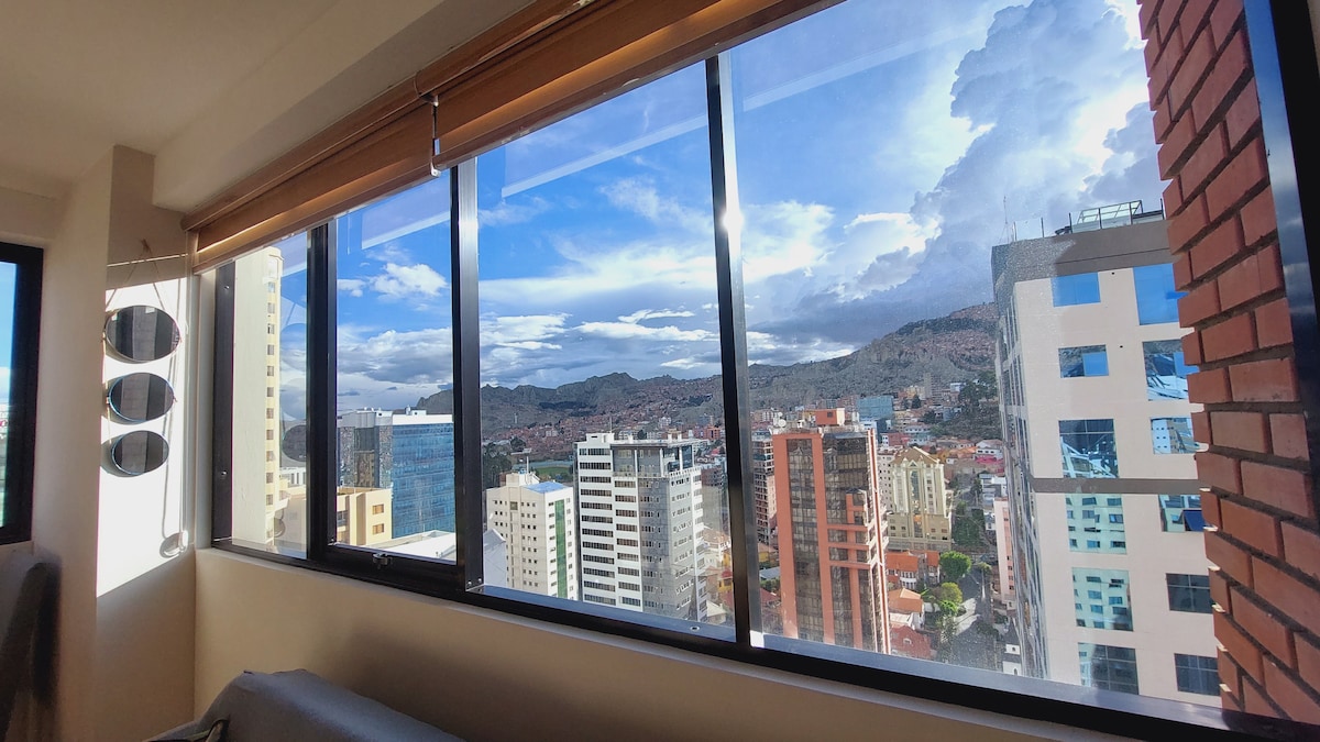 Large windows showcase panoramic views of the city and distant mountains. Natural light floods the room, enhancing the modern design with clean lines. The vibrant cityscape features a mix of buildings, greenery, and blue skies, inviting a sense of tranquility.
