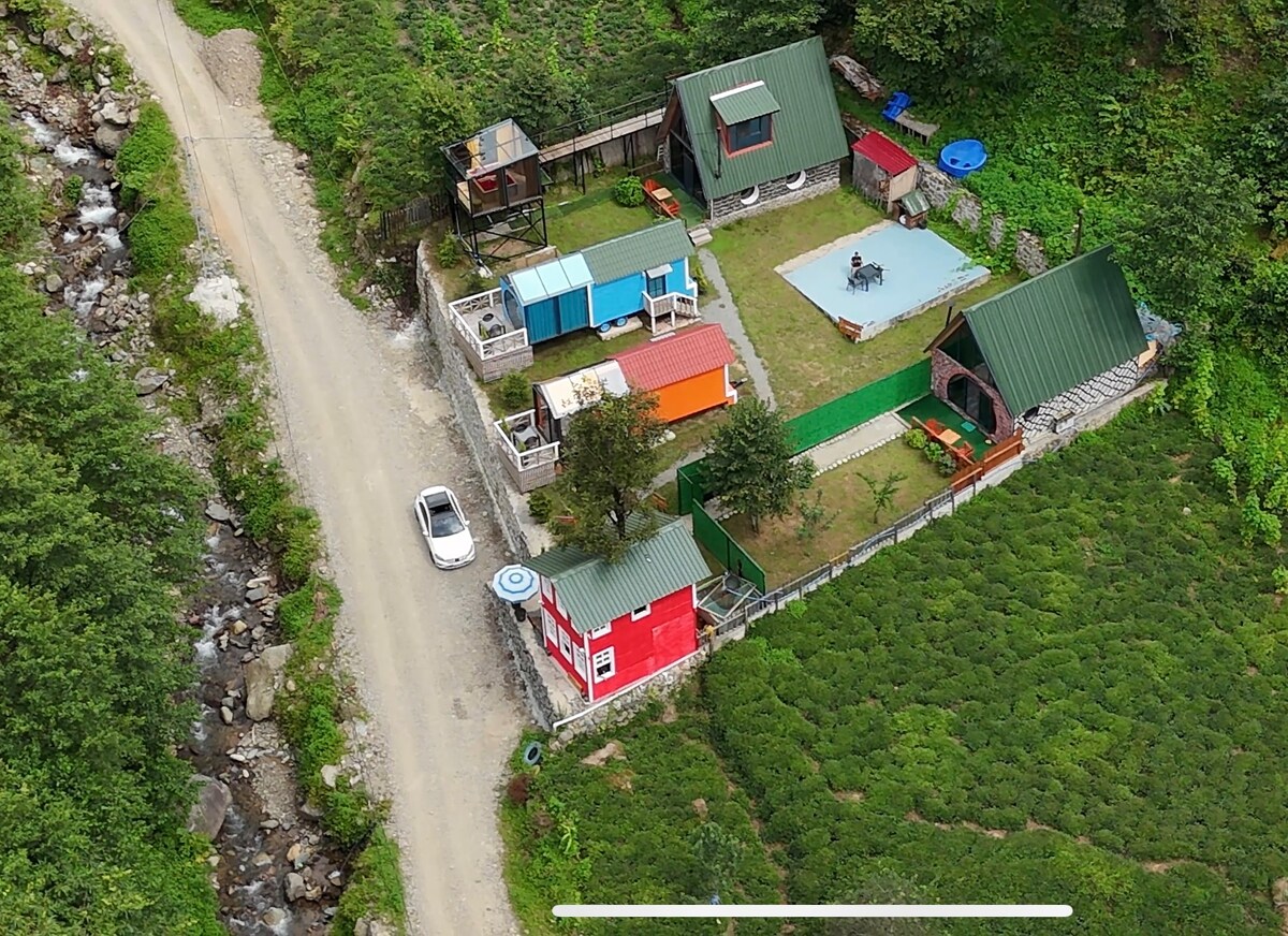 An aerial view showcases a collection of unique buildings and a swimming pool, surrounded by lush greenery. A gravel road runs alongside the property, with a car parked nearby. Outdoor furniture and shaded areas enhance the inviting outdoor space.