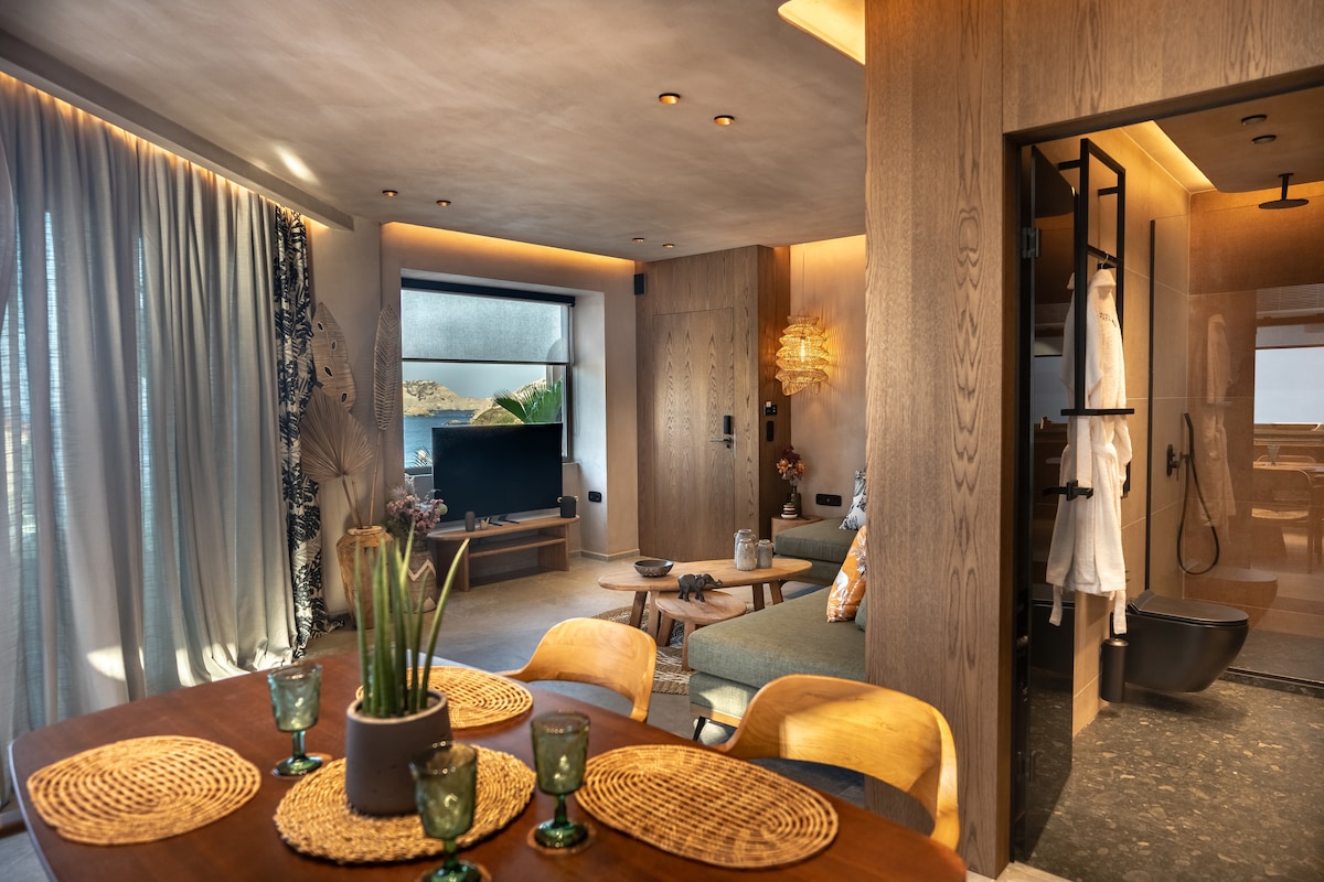 A cozy living area is complemented by a stylish dining table set for four. Soft lighting enhances the warm tones of the wooden furnishings. A contemporary television is mounted on the wall, while a spacious bathroom area is visible, showcasing modern fixtures and a soothing black bathtub.