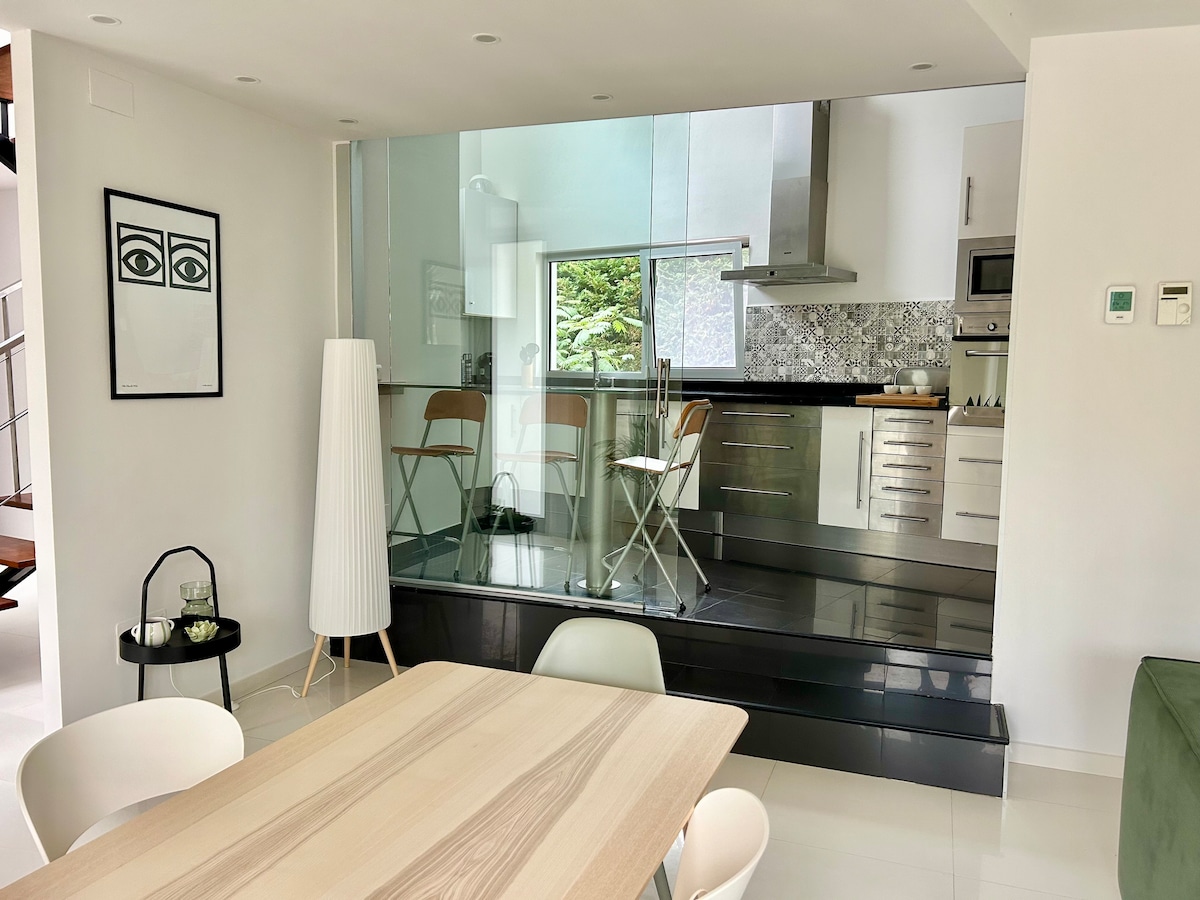 A modern kitchen and dining area are visible, featuring sleek cabinetry and stainless steel appliances. A glass partition separates the kitchen from the dining space, which includes a light wood table and white chairs. Natural light enters through a large window, enhancing the bright, open feel.