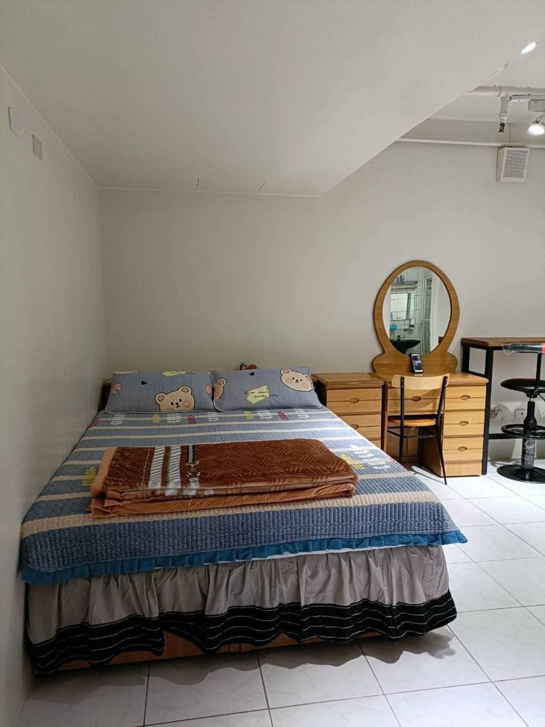A cozy sleeping area is furnished with a spacious bed topped with a patterned blanket. A wooden desk with a mirror and storage drawers is positioned nearby, alongside a tall, modern bar stool. The room features tiled flooring and neutral-colored walls.