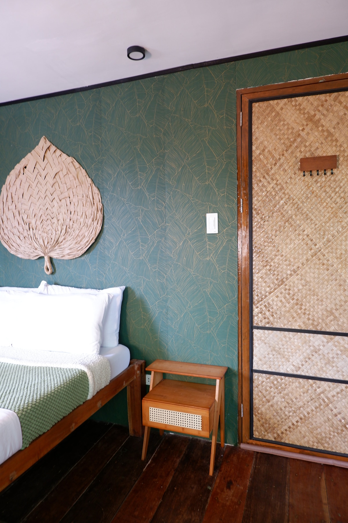 A spacious room features a rustic wooden bed adorned with crisp white linens and a green throw. A wall with leafy green wallpaper is complemented by a large woven leaf decoration. A wooden nightstand with a rattan drawer sits beside the bed, and a sliding bamboo door provides access to another area.