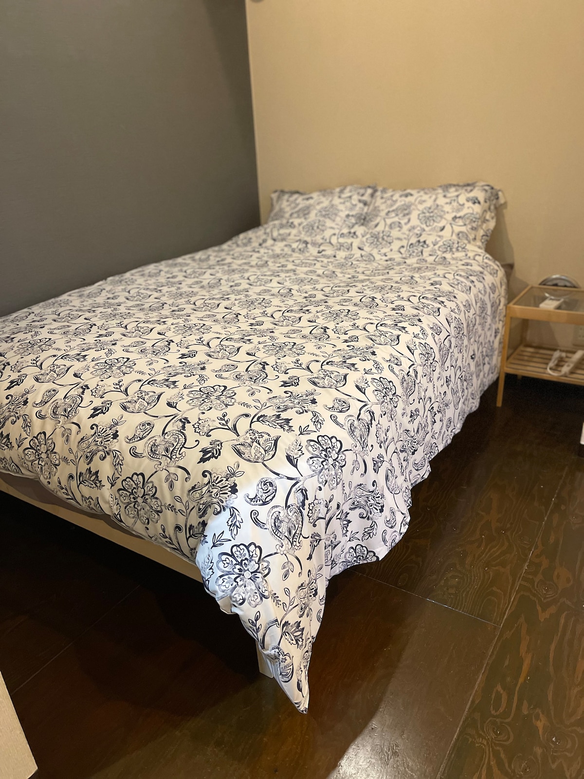 A spacious bed is adorned with a floral patterned coverlet, providing a cozy atmosphere. A small wooden nightstand is positioned beside the bed, complementing the wooden flooring and neutral-colored walls.