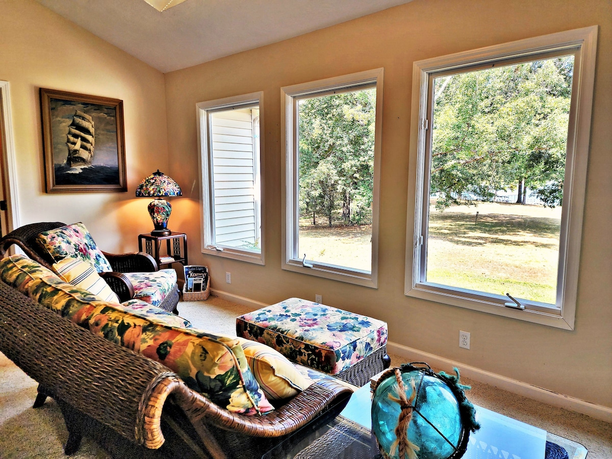 A sunlit sunroom features large windows offering views of the surrounding greenery. A wicker chair, adorned with floral upholstery, is complemented by a matching ottoman. A decorative lamp adds a touch of color, enhancing the serene atmosphere in this relaxing space.