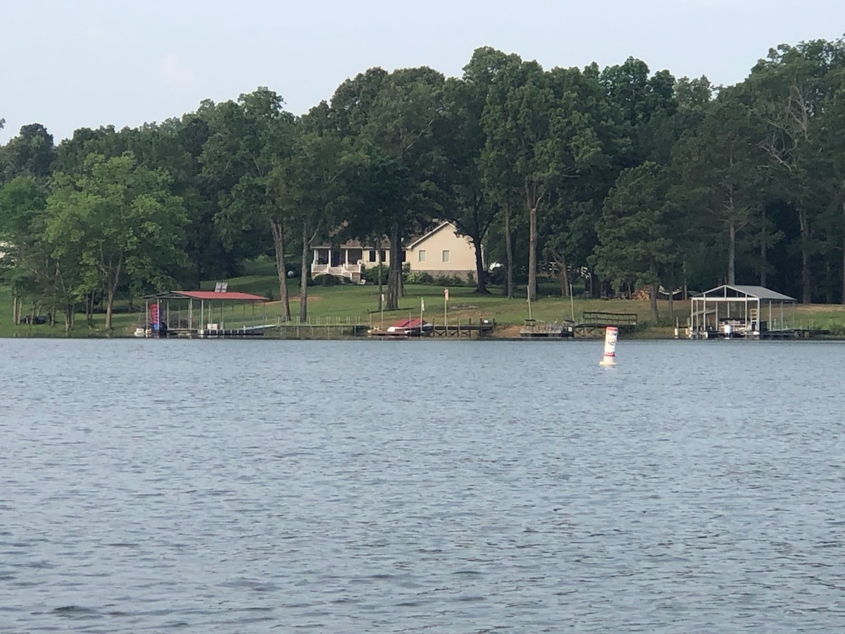 The image captures a peaceful lakeside view featuring a home nestled among trees. A private dock is visible, along with a covered boat slip. The gentle slope to the water is showcased, and the surrounding greenery enhances the serene setting.