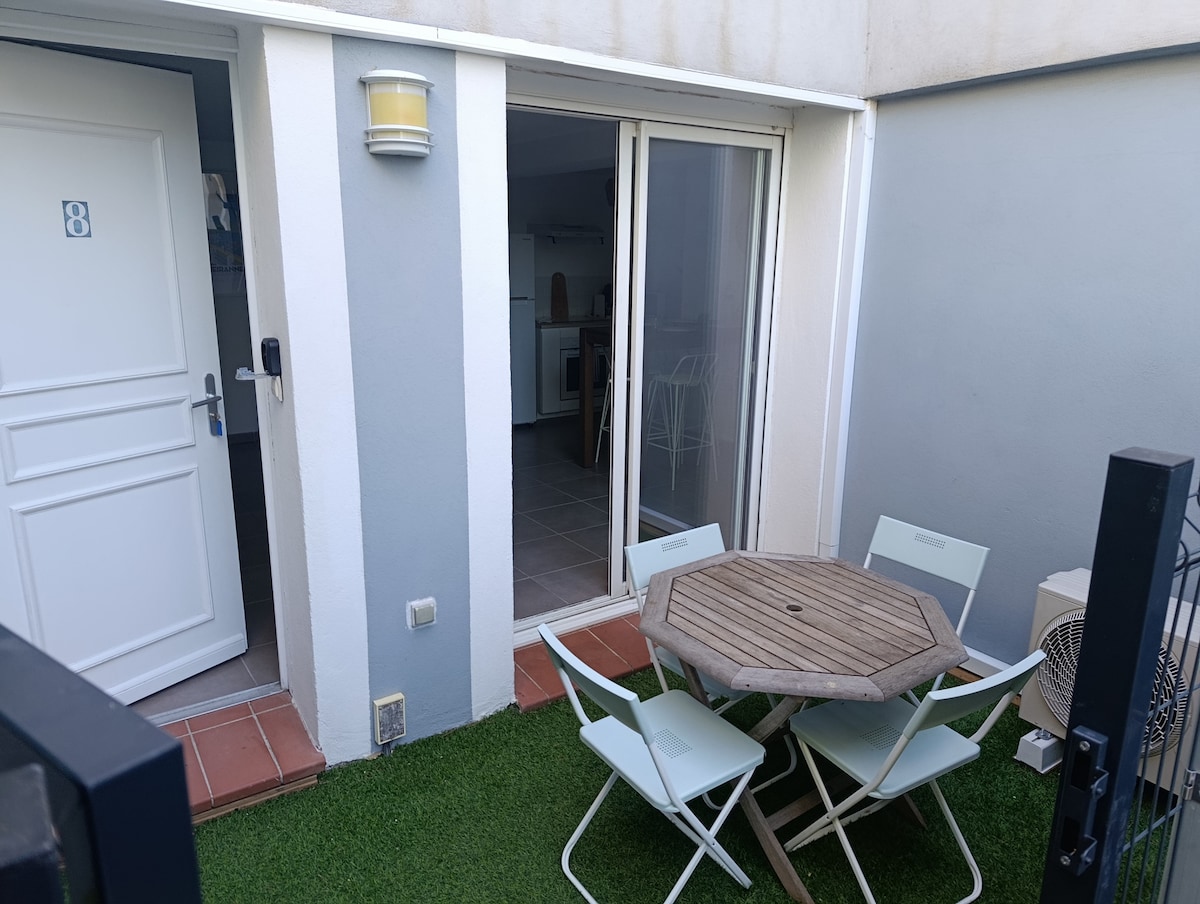 A small terrace with a grassy area features a round wooden table surrounded by four light-colored chairs. The sliding glass door opens into the living space, creating a connection between indoor and outdoor areas. A garden light is mounted on the wall adjacent to the entrance.
