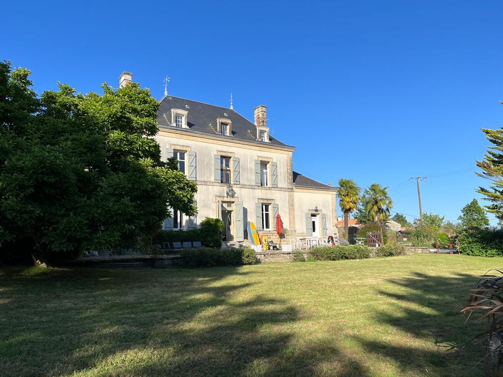 A grand house stands prominently against a clear blue sky. Lush greenery surrounds the property, and a well-maintained lawn provides ample space for outdoor activities. The façade showcases elegant architectural details, and seating areas are visible in front of the house.