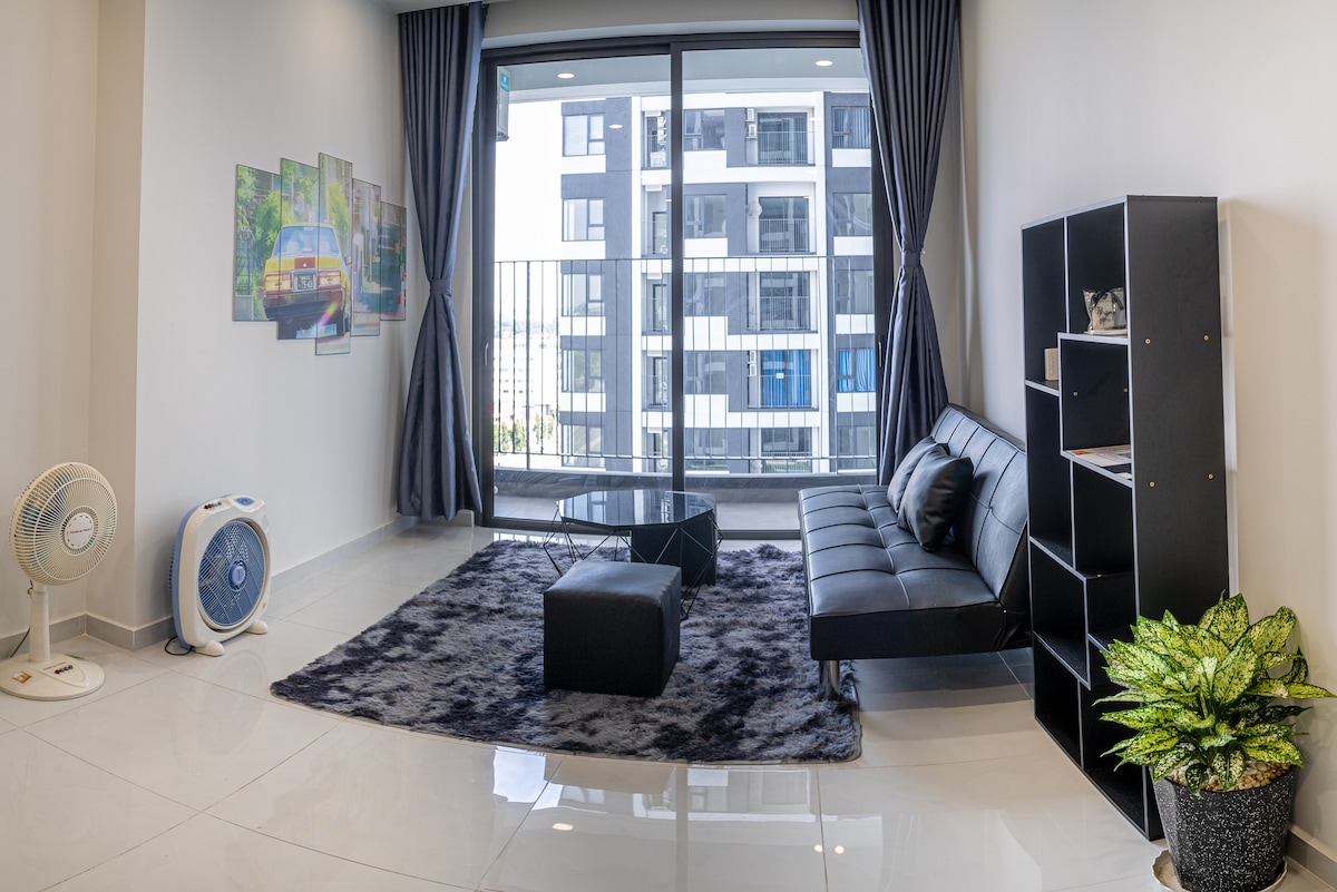 A modern living area is equipped with a sleek black sofa and a low coffee table positioned on a dark rug. Large windows offer views of the surrounding buildings, while potted plants and a decorative bookshelf provide subtle accents. A fan adds to the comfort.