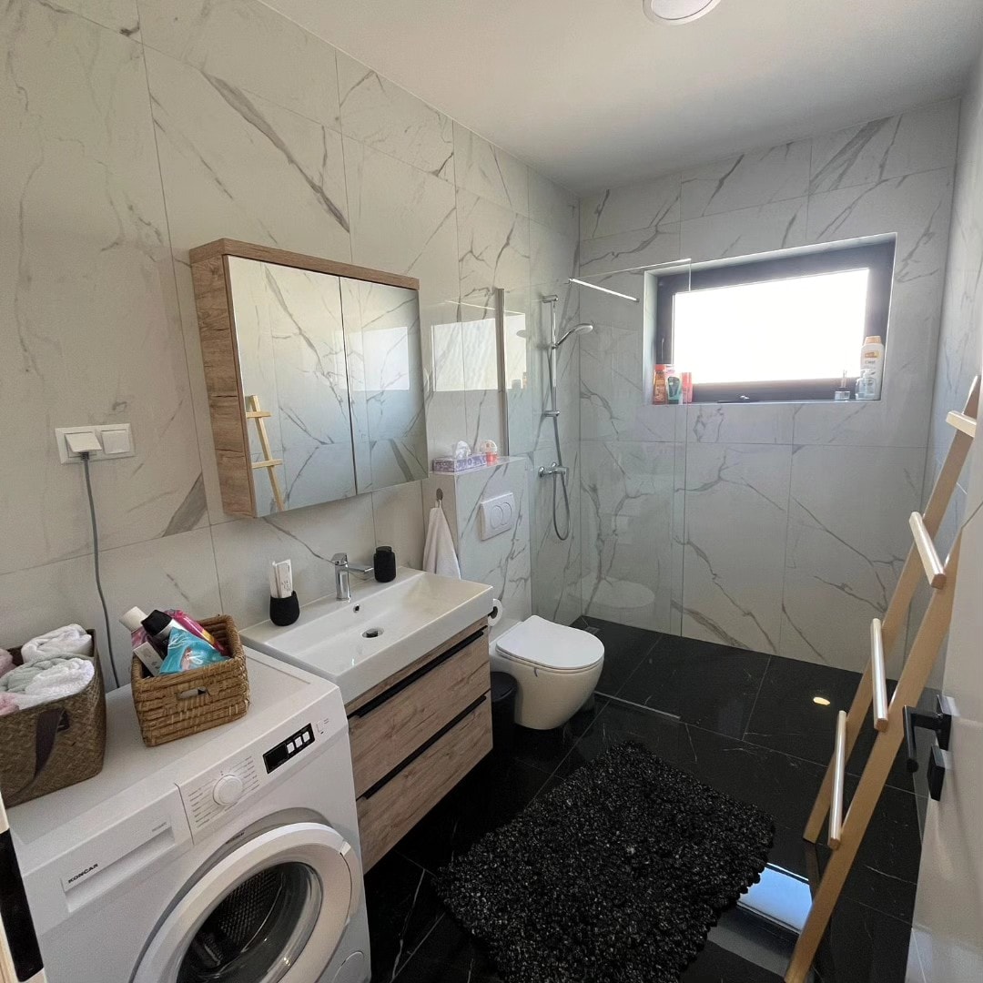The modern bathroom features a sleek design with marble-patterned walls and a dark tile floor. A washing machine is positioned next to a stylish vanity with a large mirror above. Natural light enters through a window, illuminating the space. A ladder shelf is positioned against the wall.