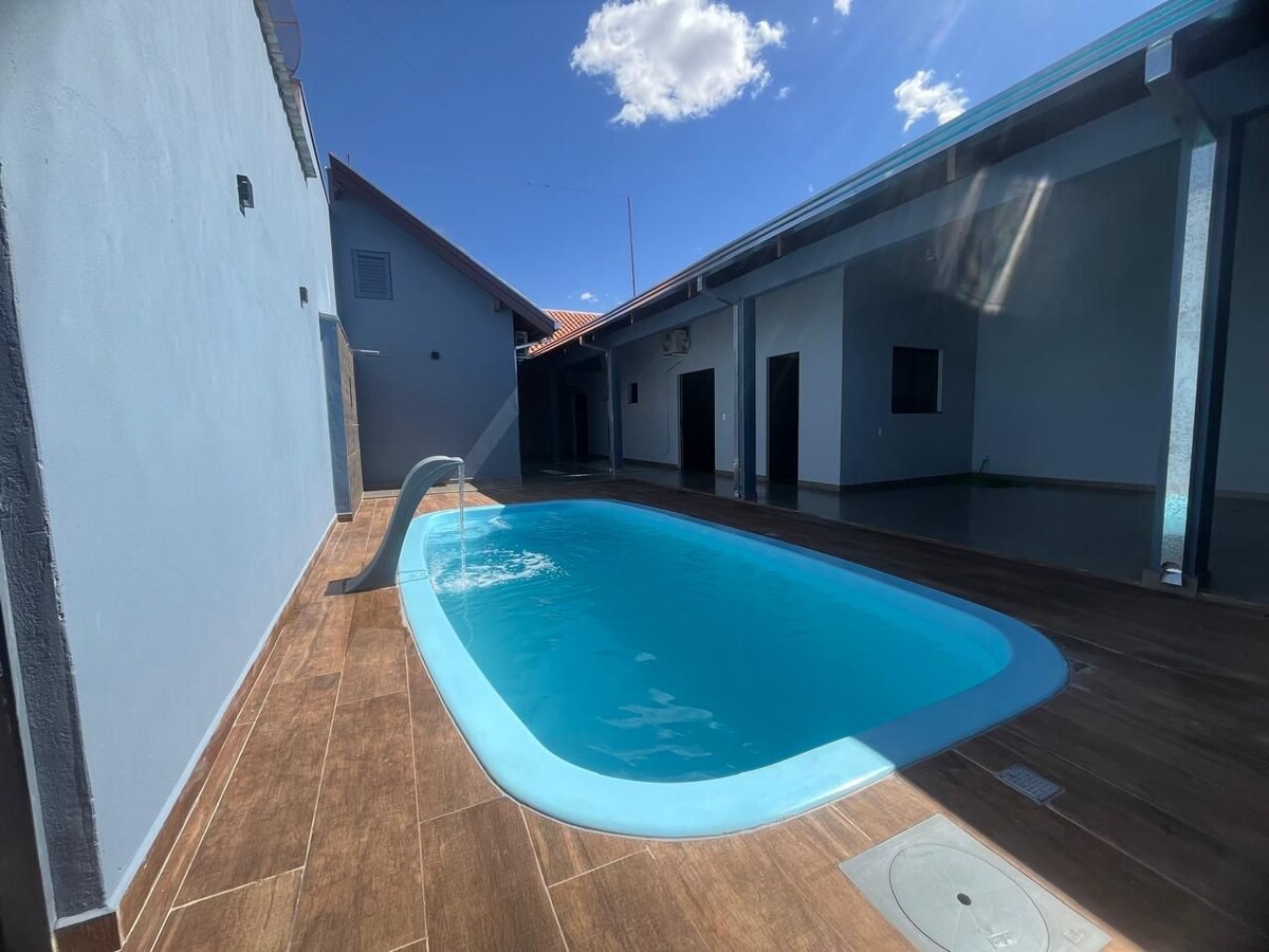 A clear blue pool is situated within a sunlit courtyard, surrounded by smooth wooden decking. The pool features a gentle slide along one side, and the area is framed by the walls of the house, creating a serene and private atmosphere.