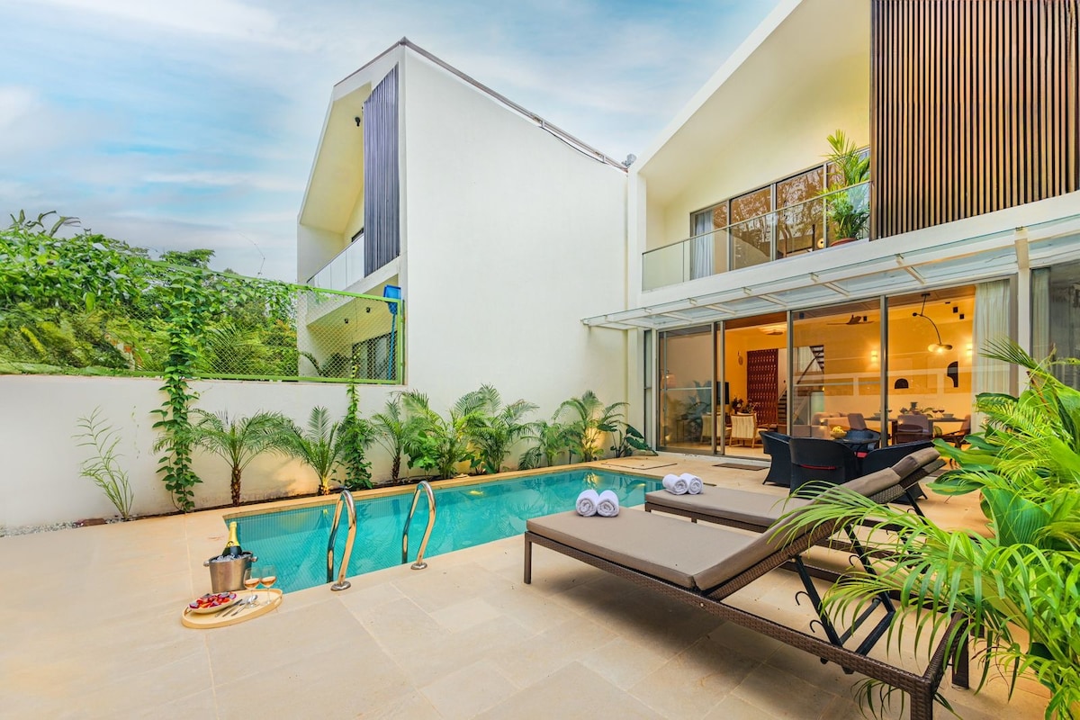 A private swimming pool is surrounded by tropical greenery, with comfortable lounge chairs positioned nearby. Glass doors provide a view into an open living space, enhancing the connection between indoor and outdoor areas. Soft lighting complements the serene atmosphere.