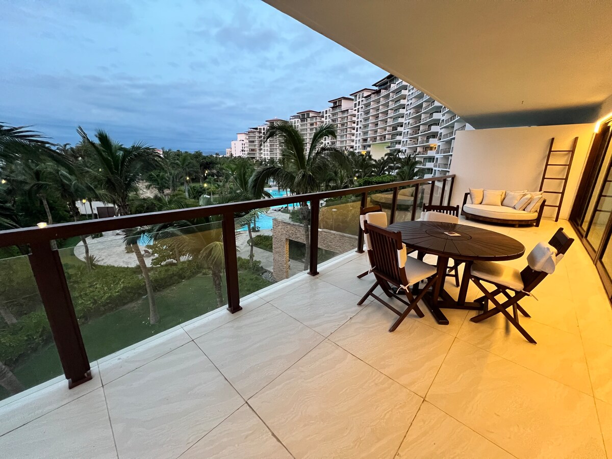 A spacious balcony features a circular wooden table surrounded by four chairs, offering a view of lush greenery and swimming pools. The area is illuminated by soft evening light, and a cozy seating arrangement with plush cushions is visible in the corner.