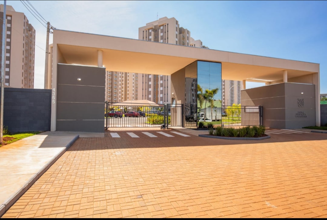 An entrance gate is visible, featuring a modern glass structure alongside a wide driveway. The surrounding area includes greenery and landscaped features, while tall buildings rise in the background, indicating a contemporary urban setting.
