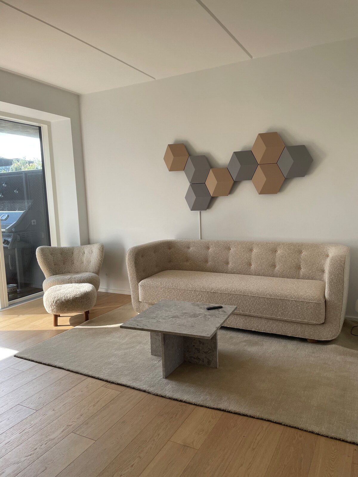 A light-filled living area features a textured, neutral sofa and a matching armchair. A geometric wall art piece adds visual interest above a simple, modern coffee table, all situated on a soft area rug that complements the warm wooden flooring.
