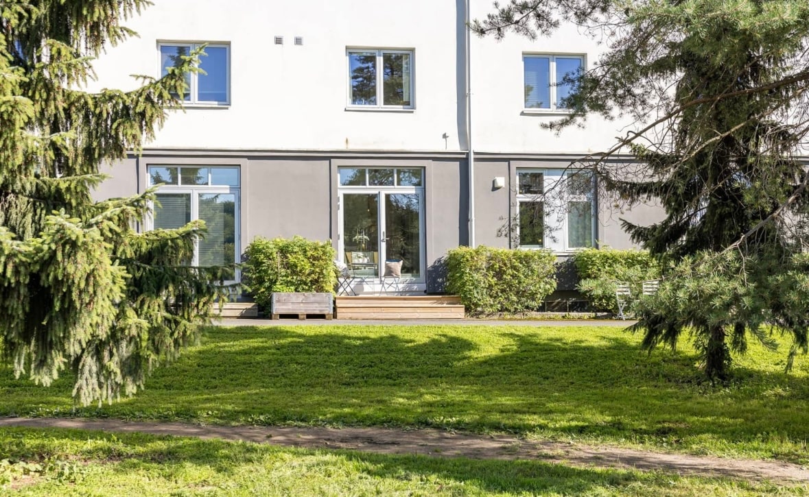 A modern building's exterior is framed by lush greenery, featuring large windows that allow natural light to enter the interior. A small deck area is visible, providing a welcoming entry point, while a well-maintained lawn extends in front of the structure, enhancing the serene environment.