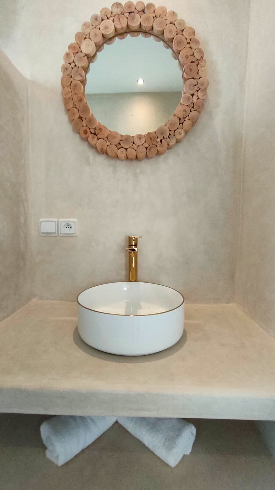A minimalist bathroom features a round, white vessel sink positioned on a smooth, beige countertop. Above, a circular mirror framed with natural wood accents complements the soft color palette. Two neatly rolled towels are placed on the countertop underneath, creating a clean and organized appearance.