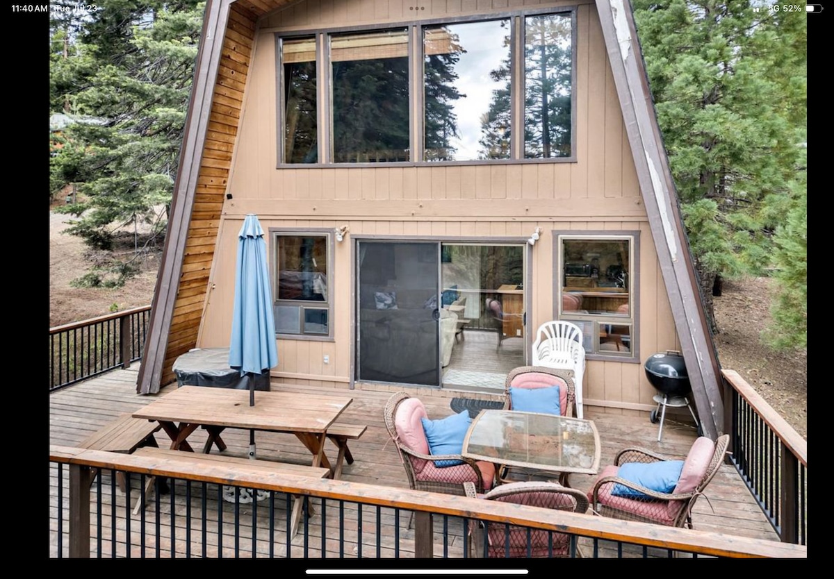 The exterior of a charming A-frame cabin is shown, featuring a spacious deck with a dining table, chairs, and an umbrella. Large windows illuminate the interior, while a grill stands nearby. The surrounding area is adorned with trees, creating a natural setting.