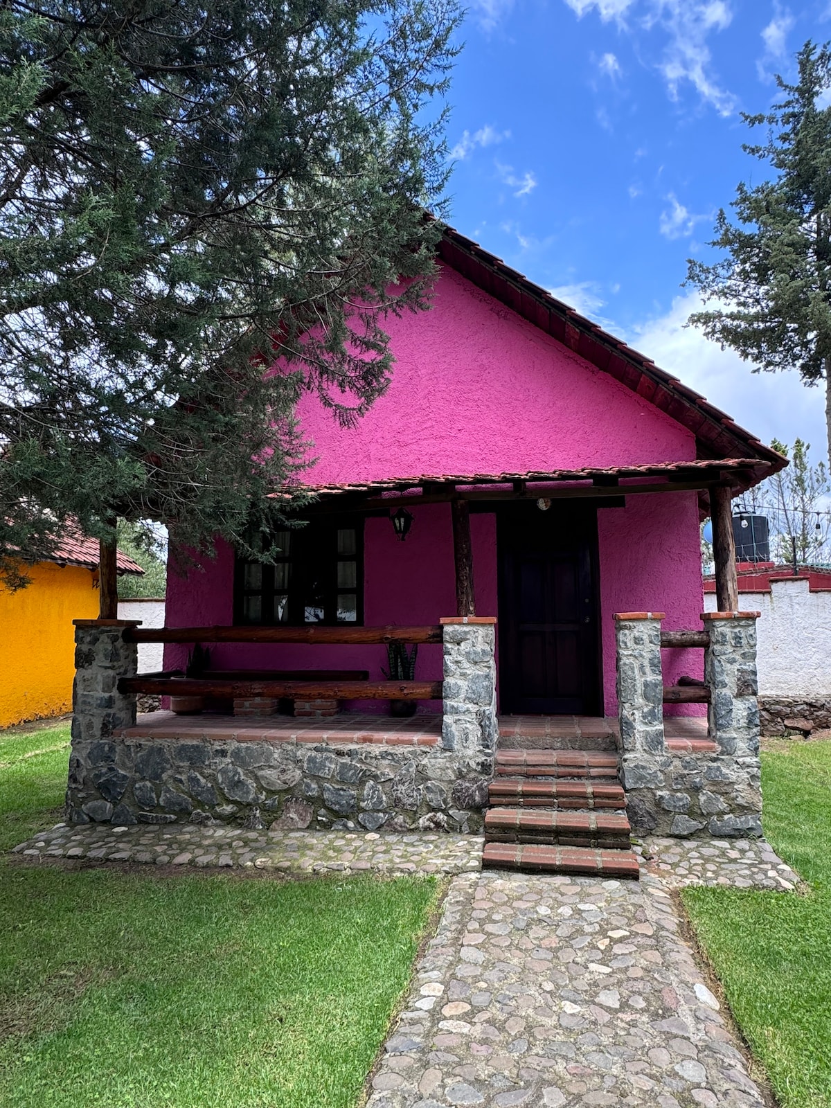 A rustic cabin with a vibrant pink exterior is framed by a stone porch. Wooden supports and a tiled roof provide architectural interest. Surrounding green grass enhances the welcoming atmosphere, while a stone pathway leads to the entrance, inviting guests to explore further.