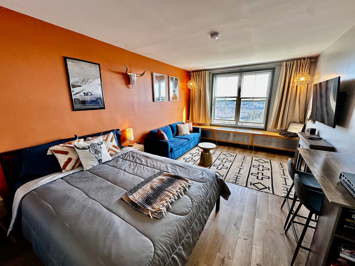 A studio space features a queen bed with layered bedding and decorative accents. A blue pull-out couch sits opposite a window with scenic views. Warm tones from the walls and lamps create a cozy ambiance. A wooden desk and two bar stools are positioned nearby.