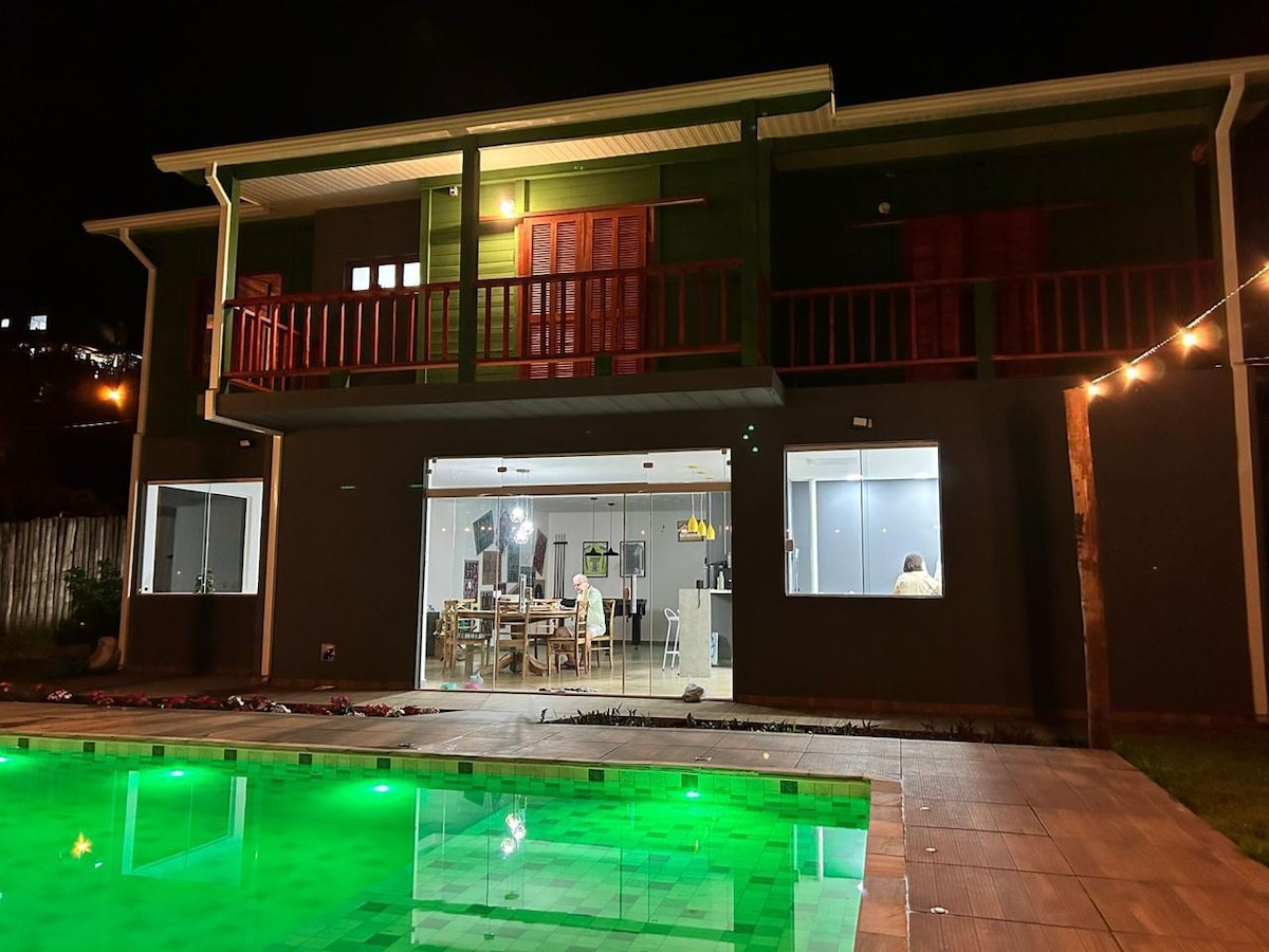 A modern two-story house is illuminated at night, showcasing large glass doors that reveal a dining area. A spacious pool glows with green light in the foreground, bordered by stone tiles. Soft lighting enhances the welcoming atmosphere around the outdoor space.