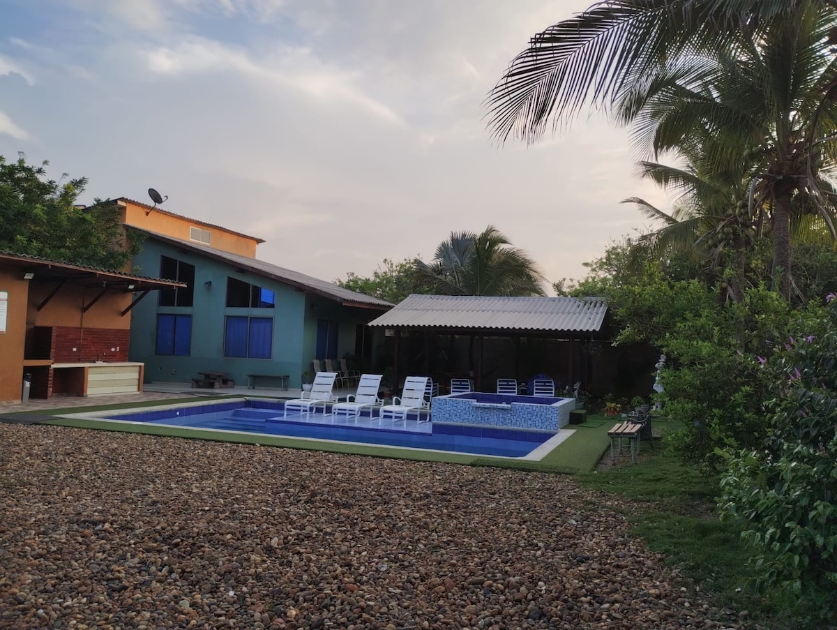 A serene outdoor area features a swimming pool surrounded by several lounge chairs. A shaded gazebo is positioned next to the pool, providing a relaxing spot. Lush greenery and palm trees are present, enhancing the tranquil setting of the property.