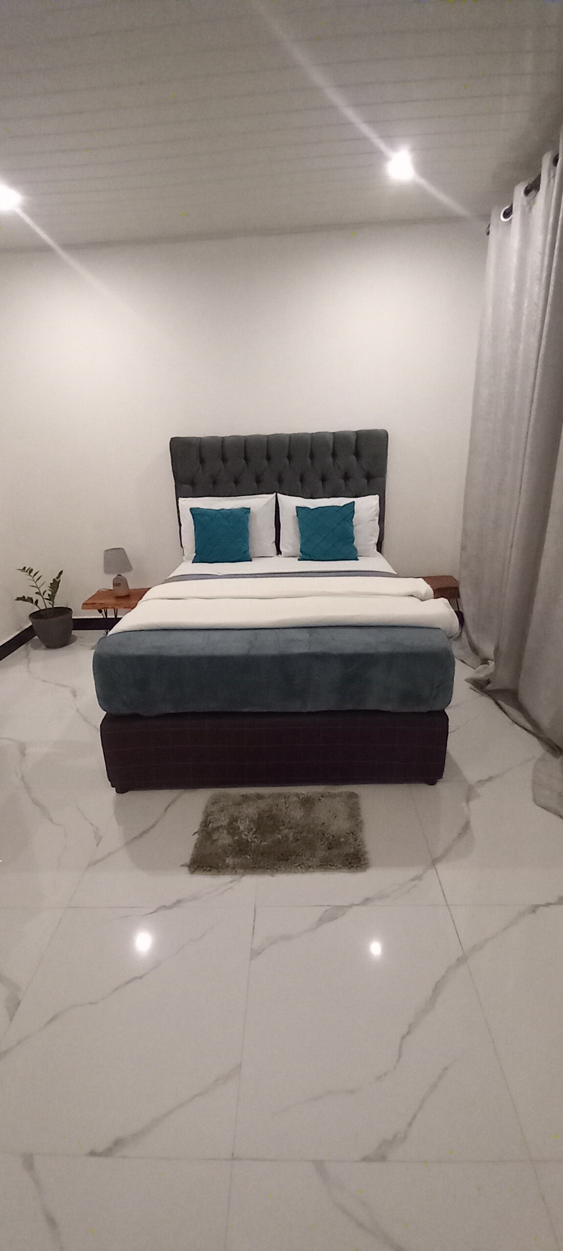 A cozy sleeping area features a tufted headboard and two decorative pillows resting on a spacious bed. A bedside table with a lamp is positioned on one side, while soft curtains frame the window. Light reflects off the smooth marble floor.