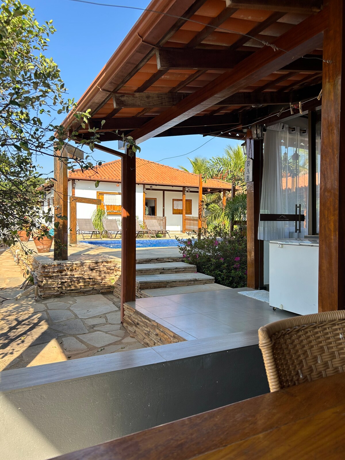 An outdoor view from a shaded porch presents a pathway leading to a pool area. Lush greenery is visible, along with a glimpse of the house featuring a stone facade and wooden accents. The clear sky suggests a sunny atmosphere.