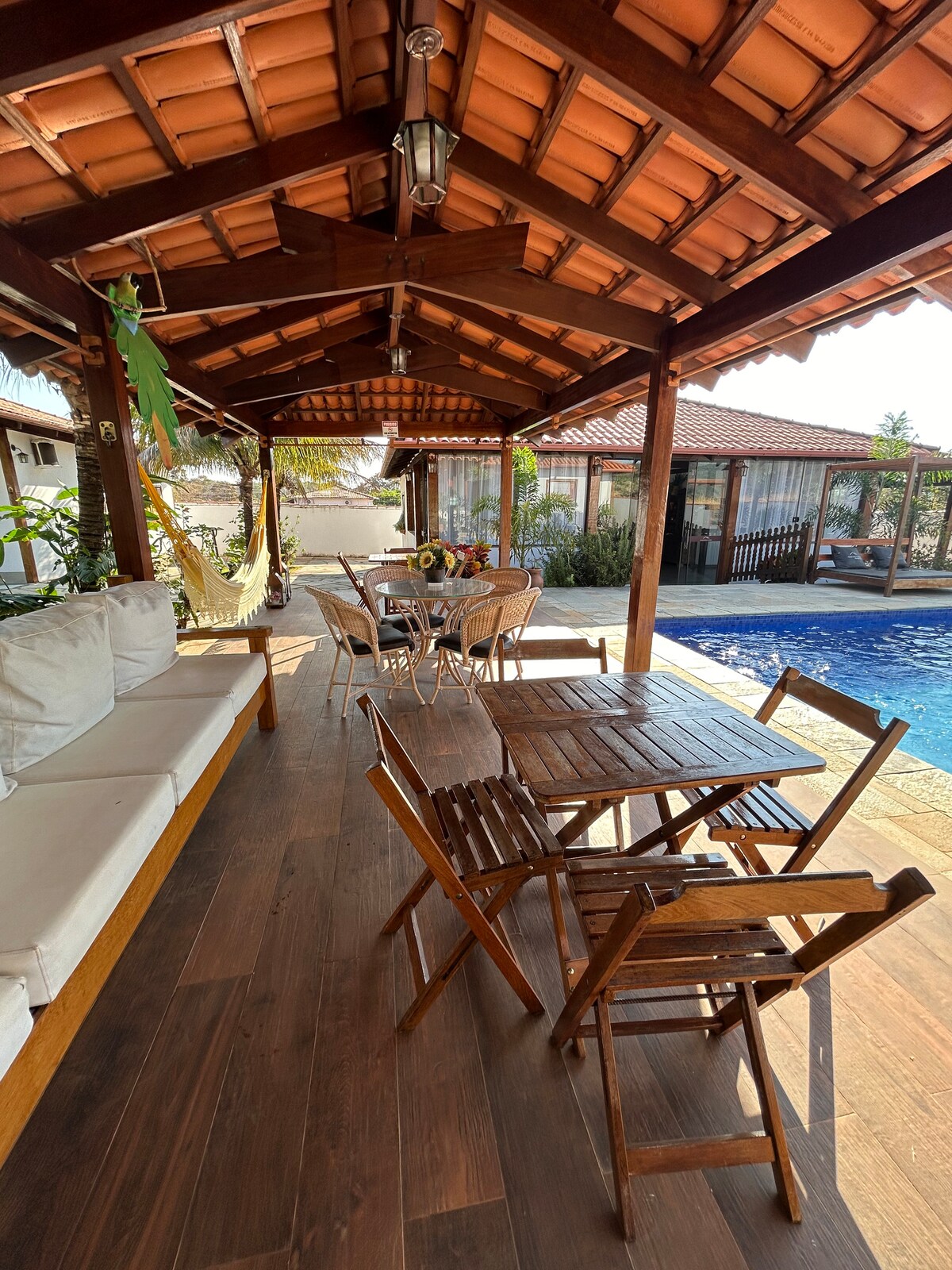 A covered outdoor seating area features a combination of a cozy sofa and a wooden dining table with chairs. Potted plants add greenery, while a hammock hangs gently nearby. The area offers a view of the pool, enhancing the peaceful setting.