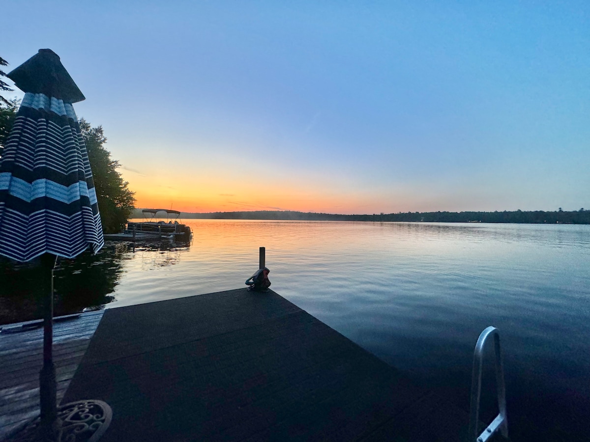 A serene lake view captured at sunset, showcasing calm waters reflecting the soft hues of orange and blue in the sky. A dock extends into the water on the left, complemented by an umbrella in a striped pattern, adding a touch of color to the tranquil scene.