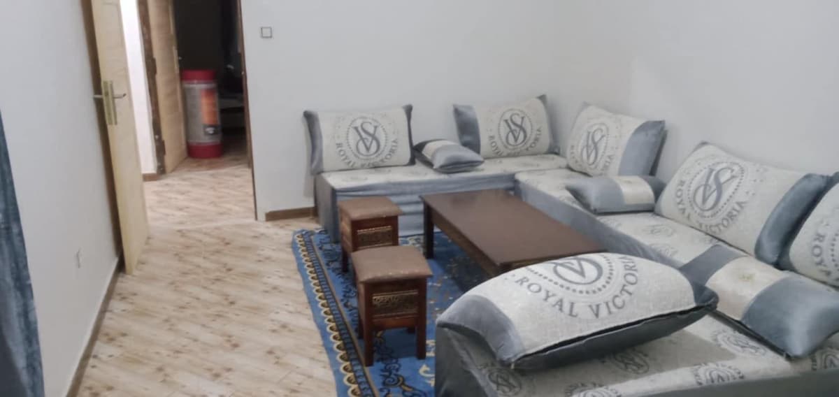 A spacious living area is furnished with a comfortable sectional sofa adorned with logo-patterned cushions. A rectangular wooden coffee table sits at the center, flanked by two small stools. A vibrant area rug adds color, while minimalistic decor contributes to a calm atmosphere.