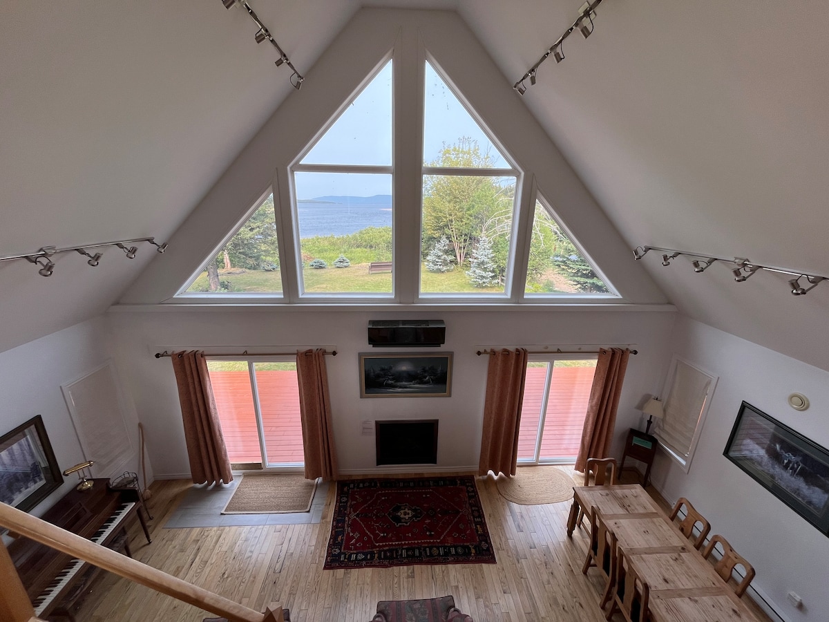 An aerial view of an A-frame chalet interior is presented, featuring a dining area with a wooden table and chairs. Large triangular windows frame scenic views of greenery and water, while light floods the space, illuminating wooden floors and wall-mounted art.