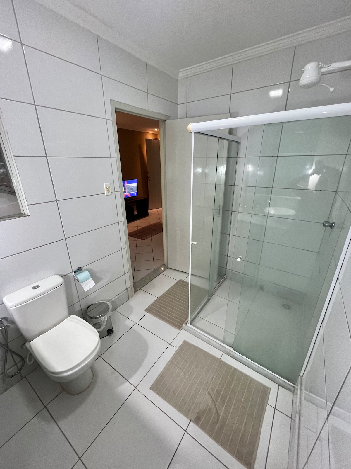 A spacious bathroom features modern white tiles and a glass shower enclosure. A toilet and a small waste bin are positioned nearby. Two bath mats provide comfort underfoot. The doorway leads to a hallway where a television is visible in the adjacent room.