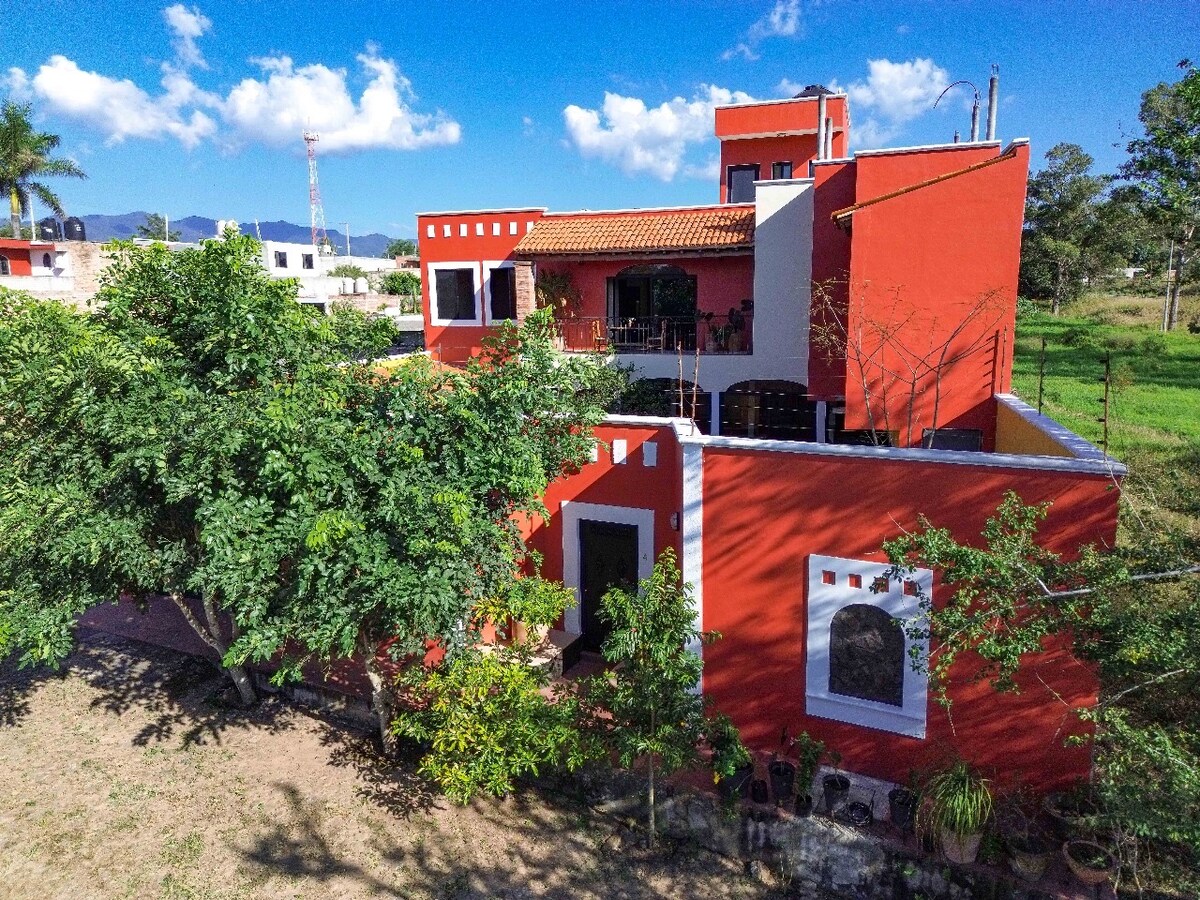 A vibrant red house is surrounded by lush greenery, featuring various windows and a spacious terrace. The structure showcases a terracotta-tiled roof and outdoor seating, offering a picturesque view of the tranquil rural environment.