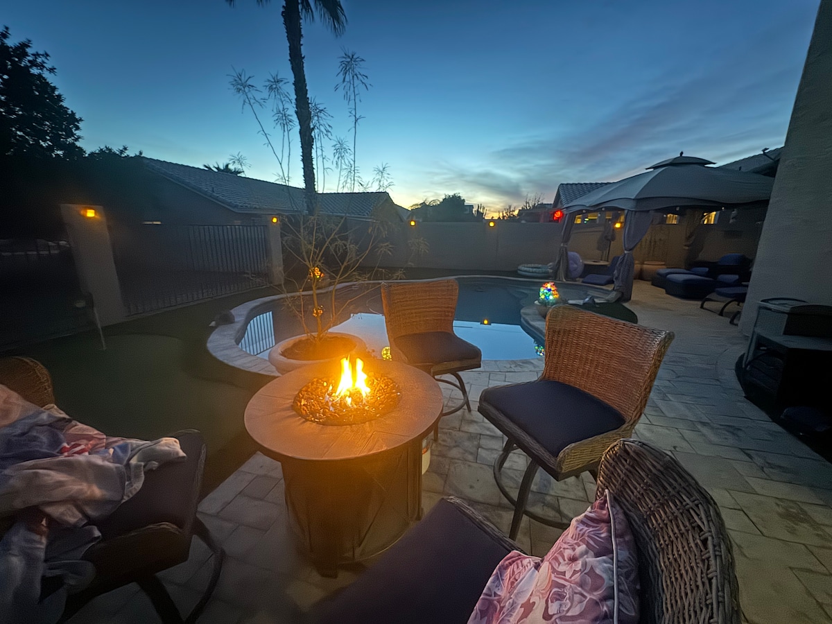 An inviting outdoor seating area features four wicker chairs arranged around a circular fire pit. The softly lit space includes a pool in the background, with palm trees and a serene twilight sky enhancing the atmosphere.