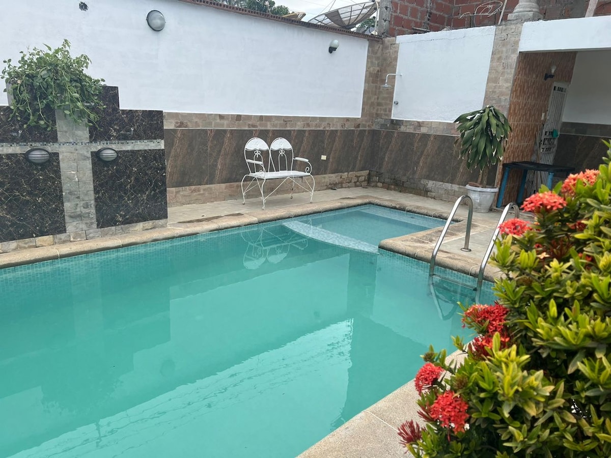 A tranquil swimming pool is surrounded by a neatly tiled area and colorful greenery. Comfortable seating, consisting of white chairs, is positioned nearby. The pool reflects the light and offers an inviting space for relaxation and leisure.