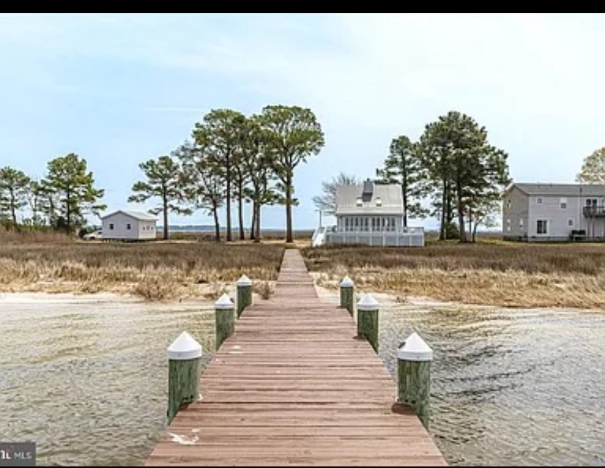A wooden boardwalk extends towards a bayfront home, bordered by tall trees and grassy areas. Numerous posts line the sides of the boardwalk, leading to tranquil water views. Two houses are visible in the background, framed by serene coastal scenery.