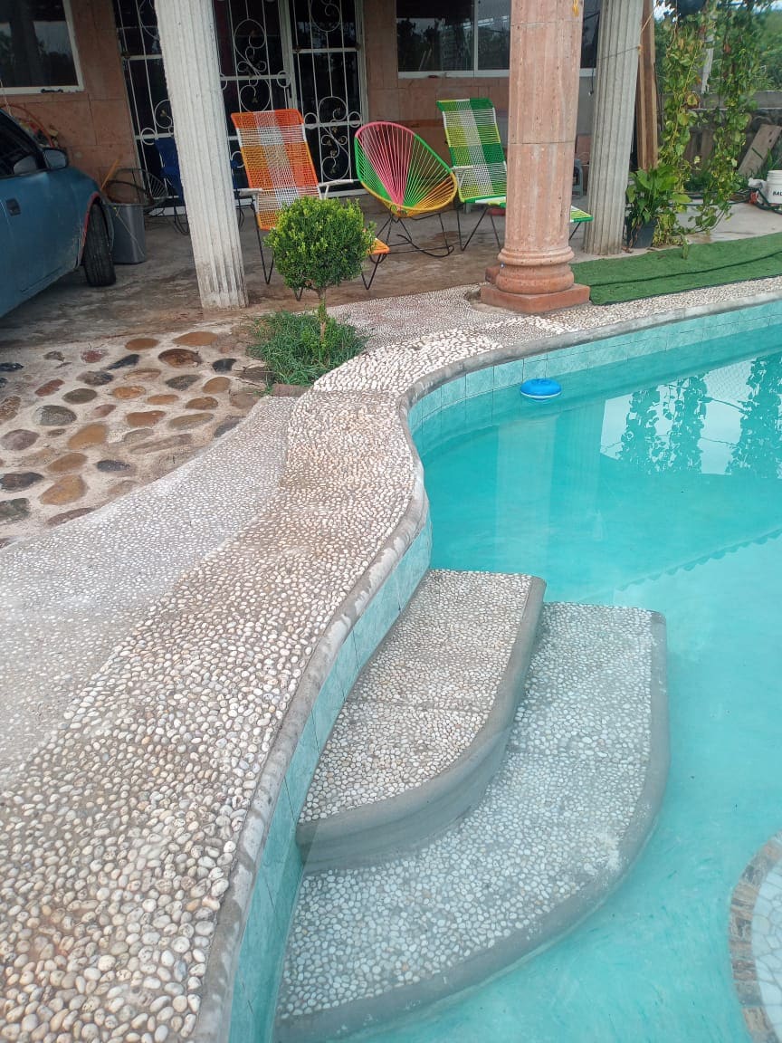 A serene swimming pool features a gently sloping entry with intricate pebble detailing. Colorful lounge chairs are positioned nearby, surrounded by greenery, creating an inviting space for relaxation.