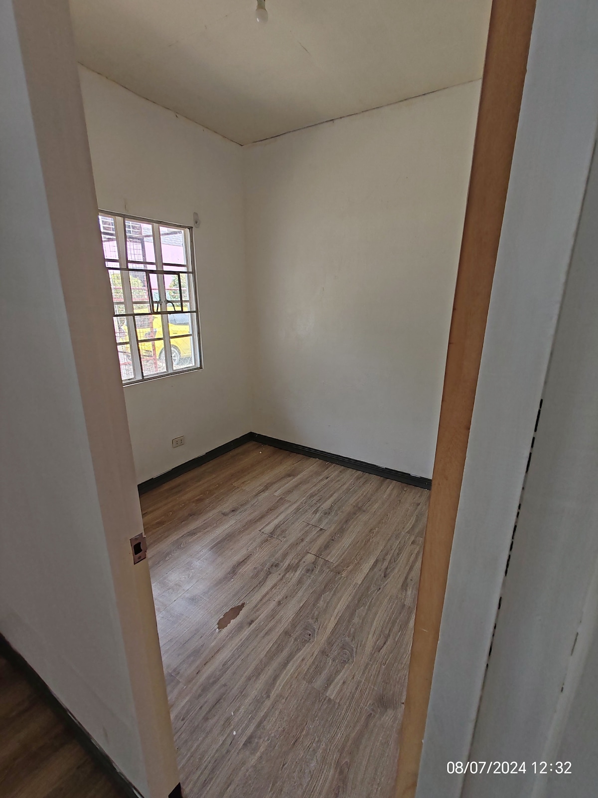 A modest room features light-colored walls and a wooden floor. Natural light enters through a window with multiple panes, adding brightness. The space remains empty, offering flexibility for various setup options.