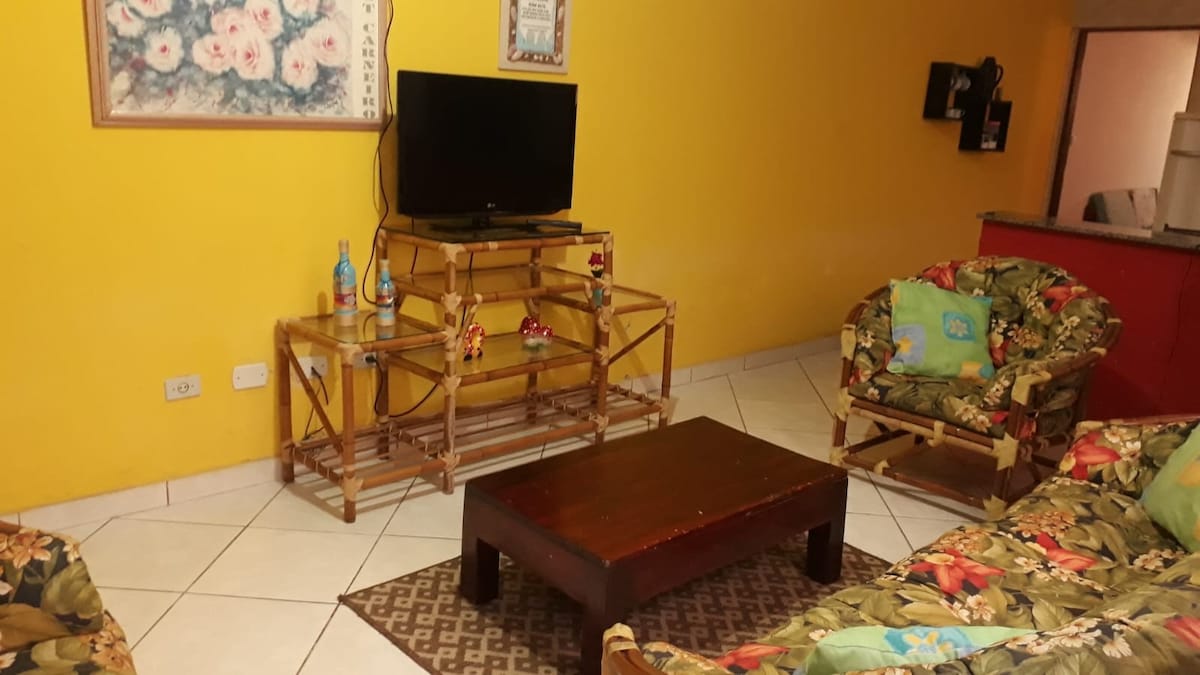 A cheerful living area is characterized by a bright yellow wall, complemented by floral-patterned seating. A low wooden coffee table sits at the center, with a TV positioned on a decorative stand. Simple decor items add a touch of color and warmth to the space.