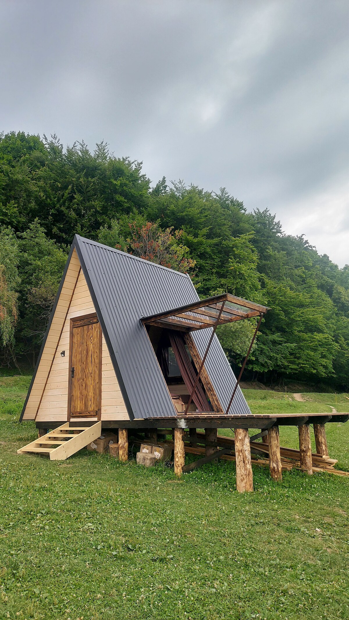 A modern A-frame cabin is set against a lush green forest backdrop. The dwelling features a unique sloped roof, large window, and a wooden deck supported by logs. Natural elements enhance the integration with the surrounding nature.
