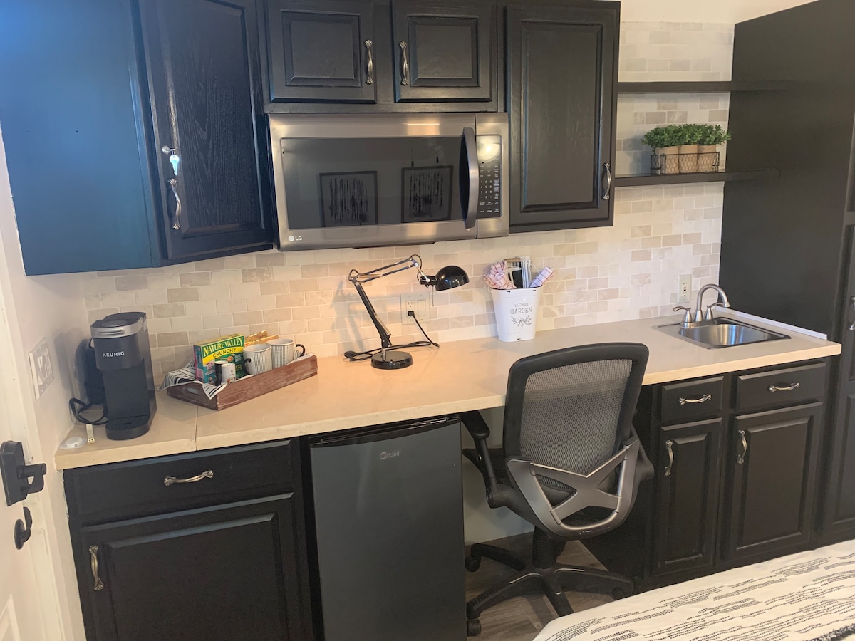 A compact kitchenette features dark cabinetry, a microwave, and a small refrigerator below the counter. A workspace includes a sleek office chair, a desk lamp, and a coffee maker, providing a functional area for work or meal preparation. Soft, neutral backsplash tiles add a clean aesthetic.