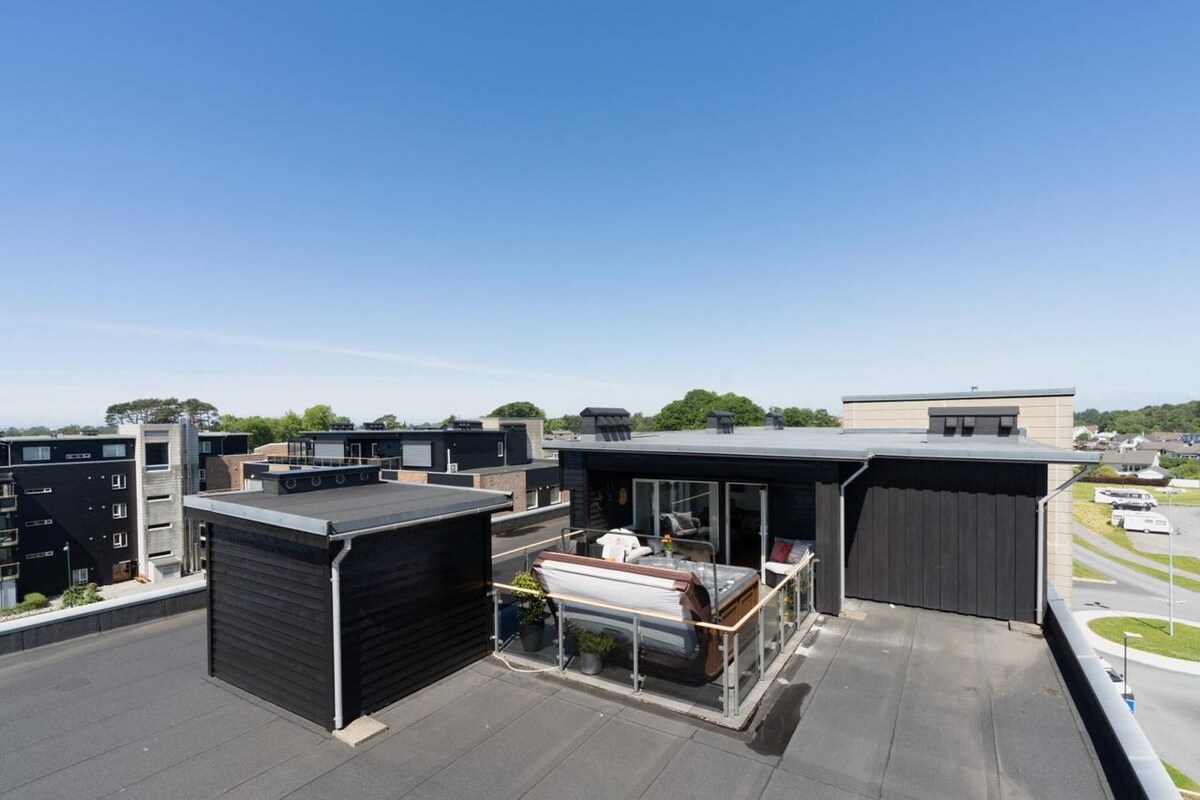 The rooftop terrace offers expansive views of the surrounding landscape, with modern black structures visible in the distance. A hot tub is positioned on a wooden deck, inviting relaxation in a sunny environment. Clear blue skies enhance the tranquil ambiance of the space.
