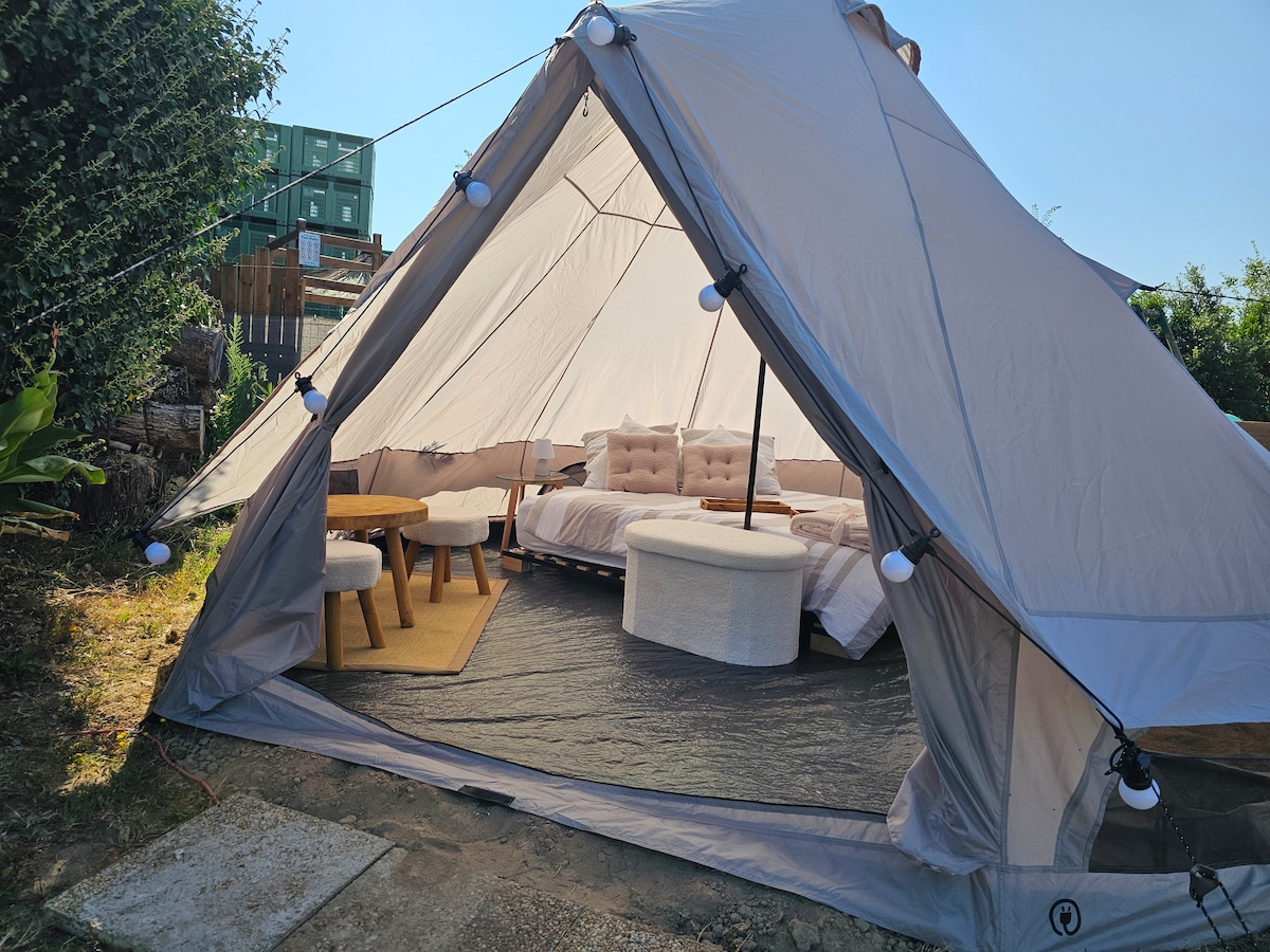 The spacious interior of the tipi features cozy furnishings, including a large bed and circular seating area. A round table is positioned near the entrance, and textured rugs enhance the comfort of the space, inviting relaxation in a serene outdoor setting.