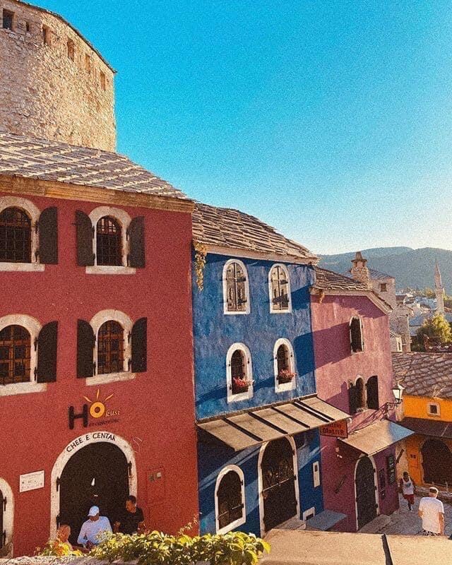 A colorful street scene features charming buildings in shades of purple and blue, accented by traditional windows and doorways. The backdrop showcases rolling hills under a clear sky, creating a vibrant atmosphere. A cafe sign is visible, inviting visitors to explore the area.