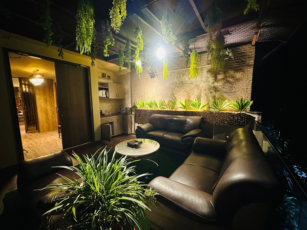 A cozy terrace garden is adorned with lush greenery, illuminated by soft lighting. Comfortable leather sofas are arranged around a circular table. The space offers an intimate setting for relaxation, with a view into the kitchen area visible through an open doorway.