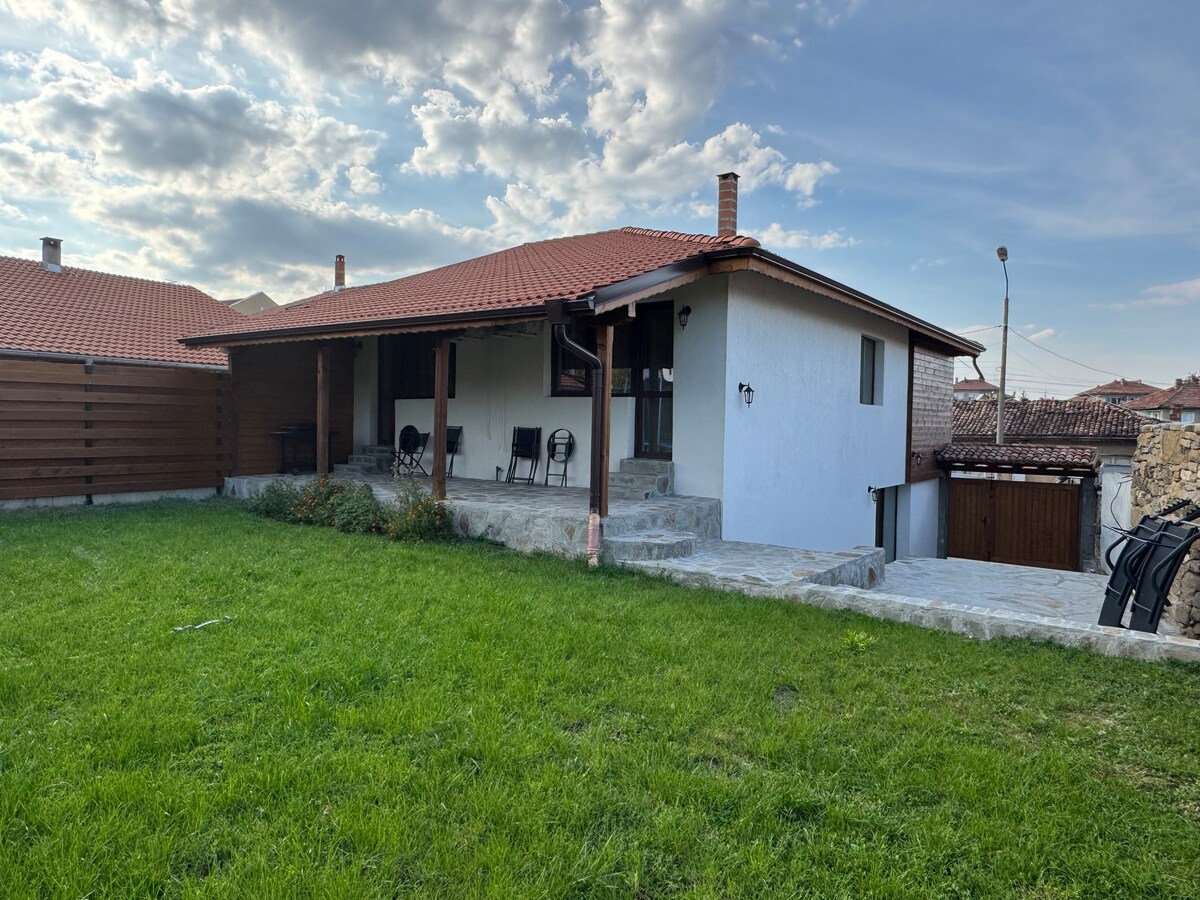 A spacious backyard is featured, showcasing a well-maintained green lawn and stone steps leading to the entrance of the house. The exterior is complemented by a traditional red-tiled roof and wooden accents, with a few chairs arranged under the covered porch.