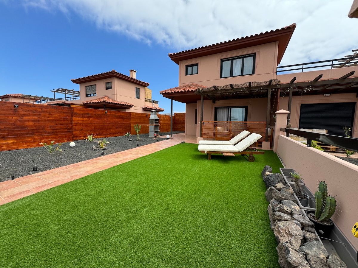 A well-maintained outdoor space features artificial turf, leading to a lounge area with two sun loungers. The home, with a rustic wooden pergola, is visible in the background alongside lush greenery and decorative plants. The area is enclosed by wooden fencing.