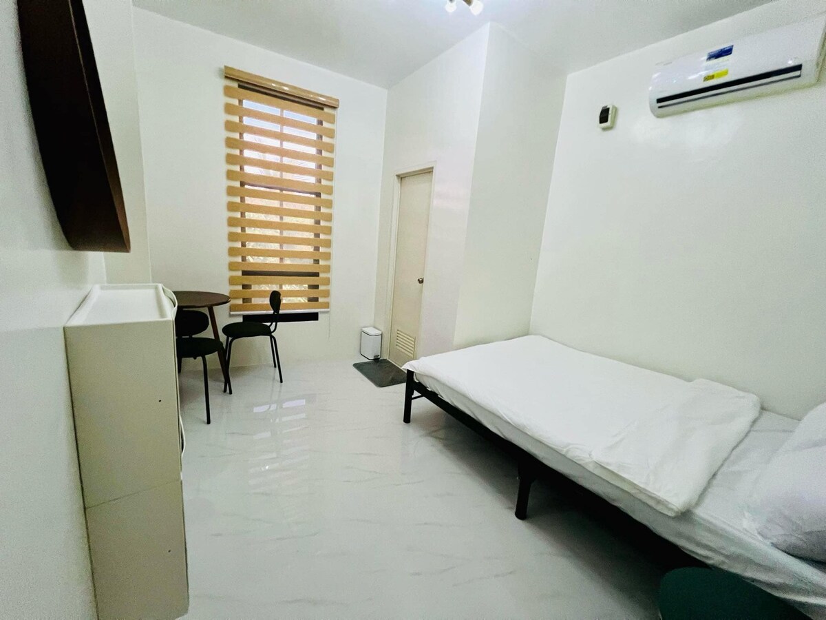 A simple studio room features a spacious bed with white linens, complemented by a small table and two chairs. Natural light filters through a honeycomb window covering, creating a bright and airy atmosphere. A modern air conditioning unit is mounted on the wall.