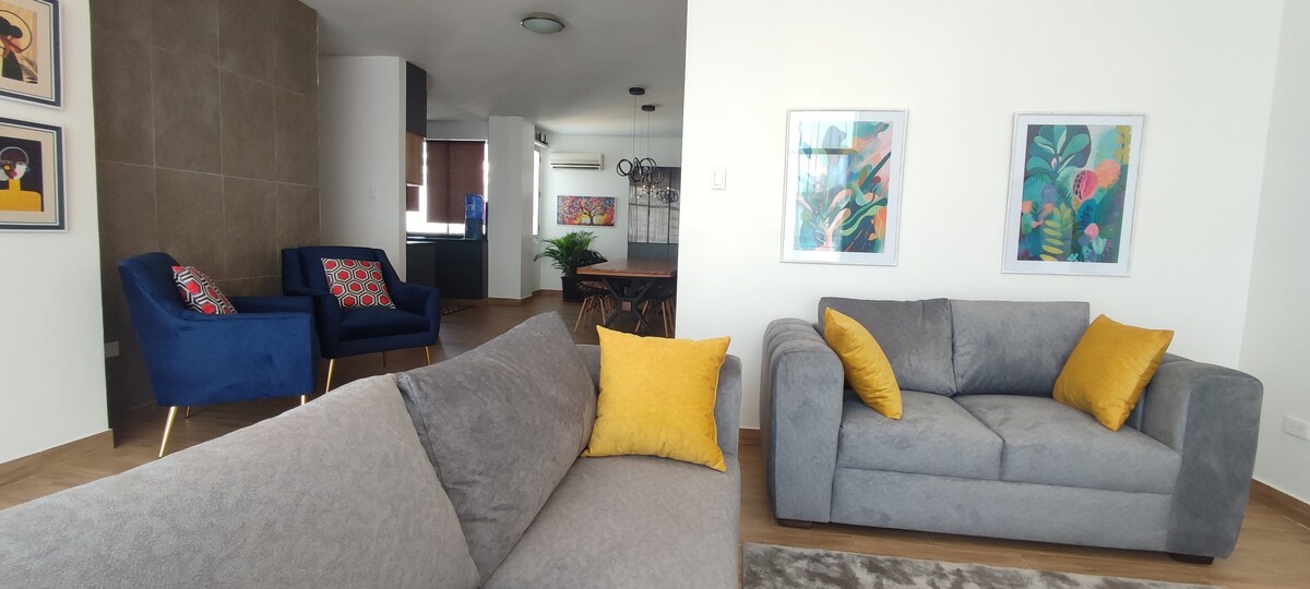 A modern living area is shown, featuring a gray sofa with yellow accent pillows and two blue armchairs adorned with colorful cushions. Framed artwork decorates the walls, and a glimpse of a dining area is visible in the background, complemented by plants and abundant natural light.